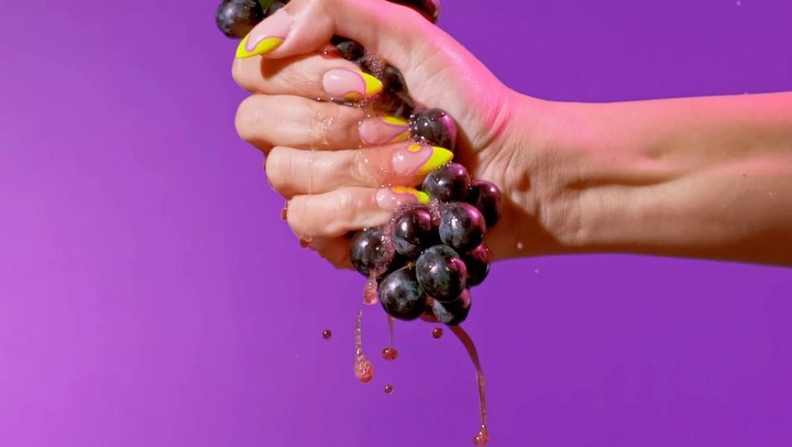Close-up of a hand with manicured nails squeezing a bunch of grapes, juice dripping against a vibrant purple background.