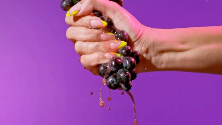 Close-up of a hand with manicured nails squeezing a bunch of grapes, juice dripping against a vibrant purple background.