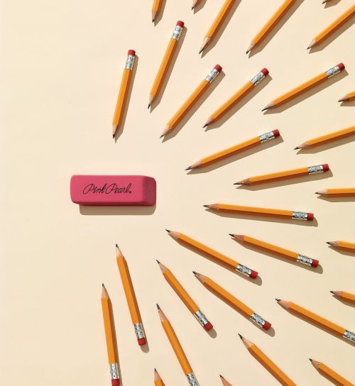 Overhead shot of a pink Pink Pearl eraser centered among several yellow pencils, all arranged on a light beige background for a school or office theme.