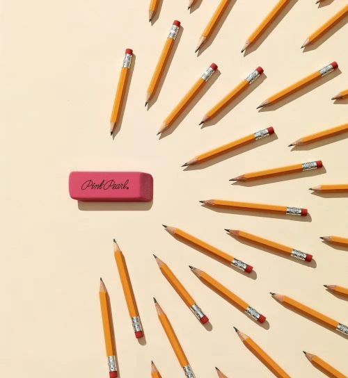 Overhead shot of a pink Pink Pearl eraser centered among several yellow pencils, all arranged on a light beige background for a school or office theme.