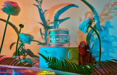 GlamGlow Moisturetrip product shot on a turquoise block with flower decorations and warm-toned lighting in a studio setting.