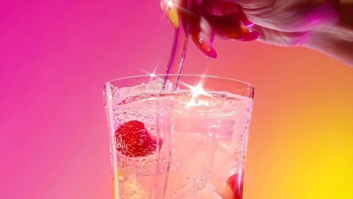Close-up of a hand pouring a clear liquid into a glass of ice and strawberries with a pink and yellow gradient background.