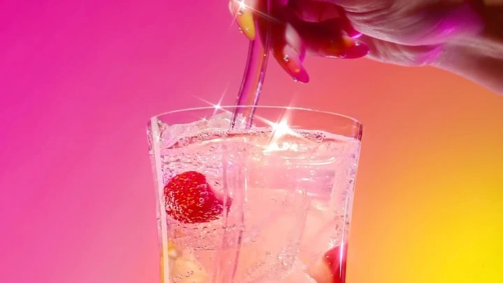Close-up of a hand pouring a clear liquid into a glass of ice and strawberries with a pink and yellow gradient background.