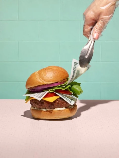 Gloved hand places 100 dollar bills inside a hamburger with lettuce, tomato, onion on a pink table against a teal tile wall.