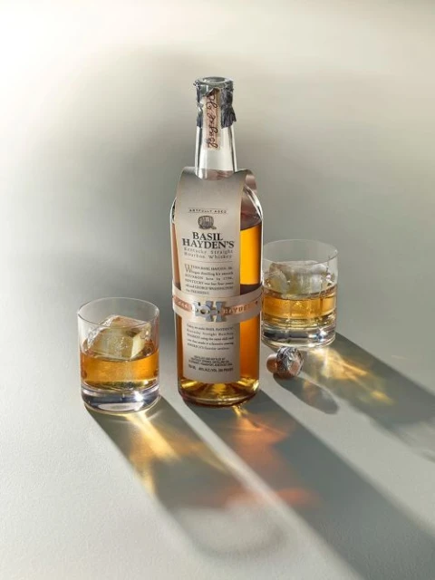 A bottle of Basil Hayden's bourbon whiskey is featured with two glasses of whiskey and ice on a white surface in a studio setting.