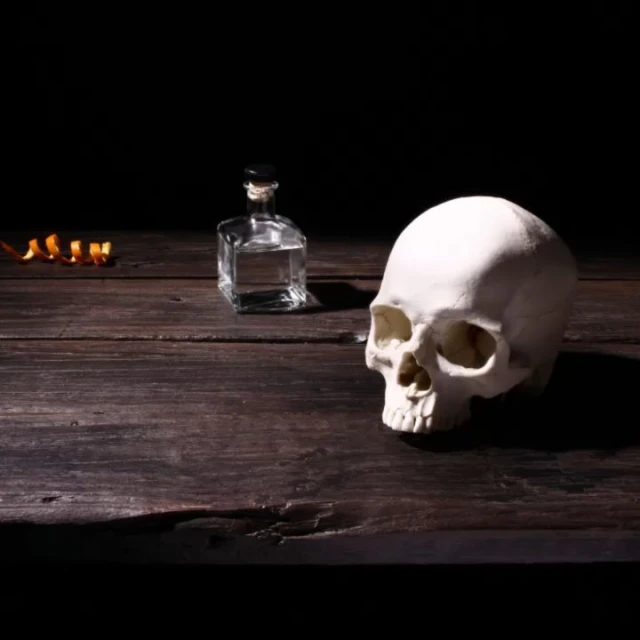 Still life with skull, a glass bottle, and an orange peel arranged on a wooden table against a dark background.
