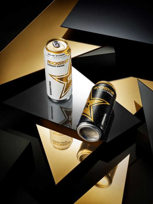 Two Rockstar energy drinks are displayed with reflections and gold/black geometric shapes in the background, creating a dynamic composition.