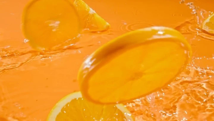 Slices of oranges are shown suspended in mid-air, splashing into a pool of vibrant orange juice, capturing a moment of freshness and movement.