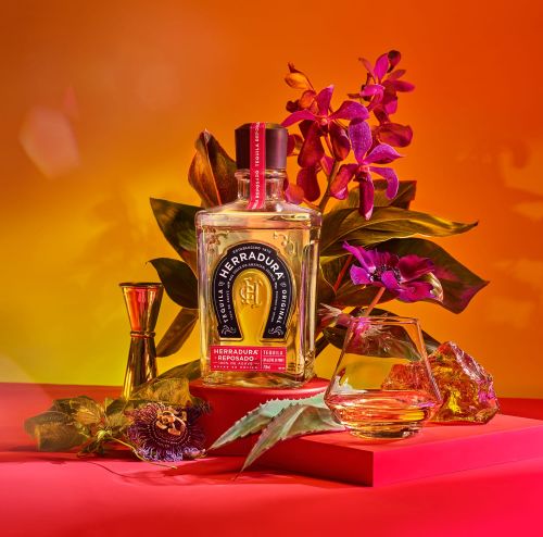 Studio shot of a bottle of Herradura tequila, a glass of tequila, and a jigger, staged with flowers on a red platform against a gradient background.
