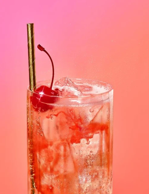 Tall glass filled with bubbly pink beverage, ice, gold straw, and a bright red cherry against a vibrant gradient background.