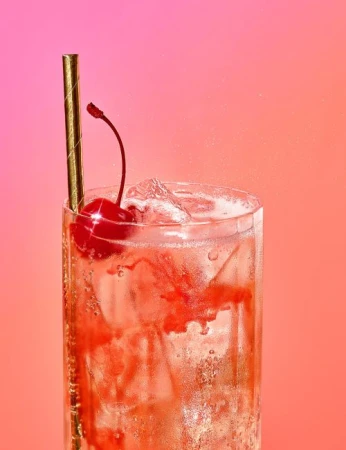 Tall glass filled with bubbly pink beverage, ice, gold straw, and a bright red cherry against a vibrant gradient background.