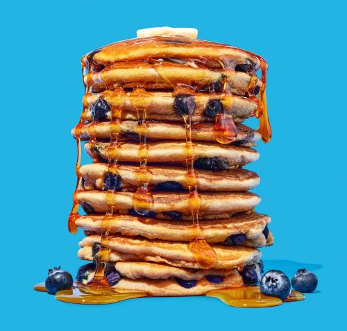 Tower of blueberry pancakes with melting butter and syrup on a blue background. Food photography showcasing a breakfast item.