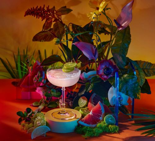Cocktail in coupe glass garnished with lime, surrounded by flowers and fruit slices in a vibrant, colorful arrangement.
