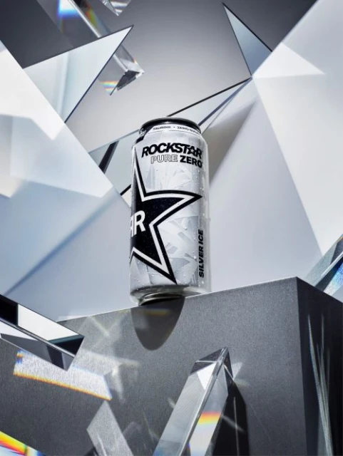 A single can of Rockstar Pure Zero Silver Ice energy drink sits on a gray platform against a background of mirrored shards.