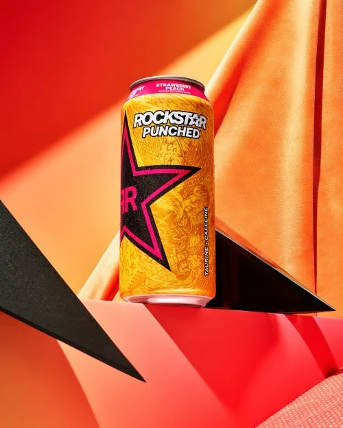 A can of Rockstar Punched Strawberry Peach energy drink is showcased against a vibrant backdrop of orange and black geometric shapes.