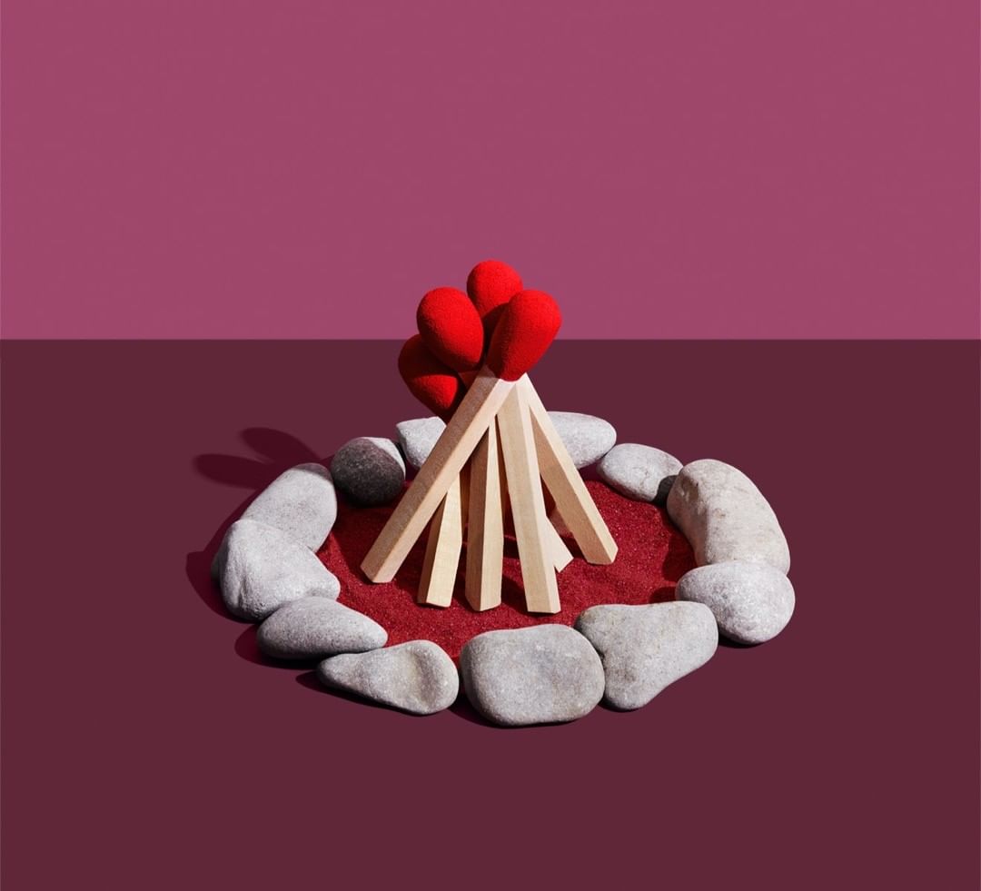 Campfire built from wooden sticks, red heart shapes, and grey stones on a purple background in a stylized still life.