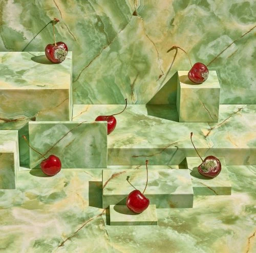 Studio shot of cherries with stems, some with mold, arranged on geometric platforms in a green marbled pattern, creating an artistic food still life.