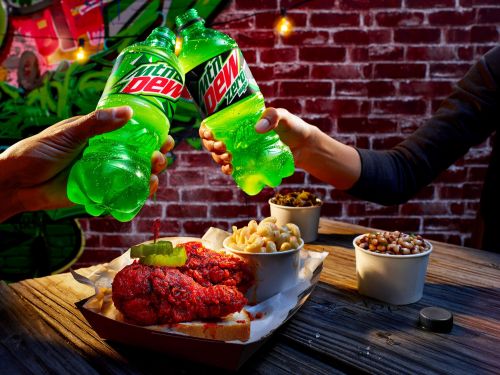 Two people toasting with bright green Mountain Dew bottles, food on a wooden table, brick wall background, and colorful graffiti art.