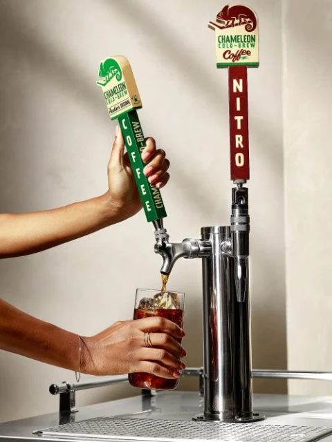 Hands pour Chameleon Cold-Brew coffee from a dispenser into a glass with ice. Dual-tap system advertises nitro coffee variant.