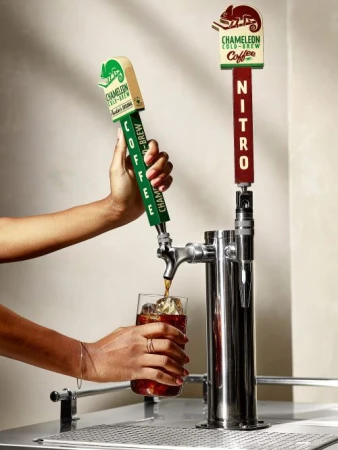 Hands pour Chameleon Cold-Brew coffee from a dispenser into a glass with ice. Dual-tap system advertises nitro coffee variant.