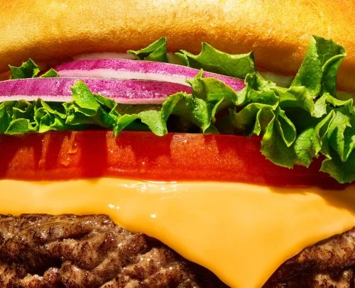 Close-up of a juicy hamburger with a beef patty, melted cheese, tomato, lettuce, and red onion on a toasted bun.