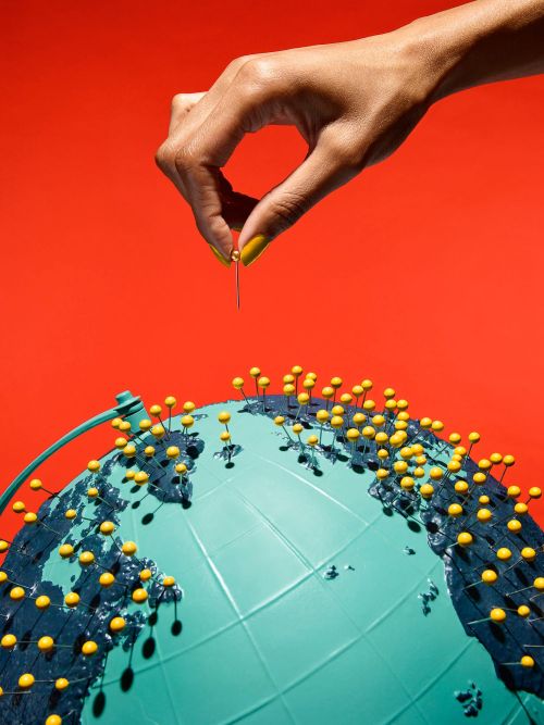Human hand using pin to mark a blue globe with yellow pinheads on a red background, visualizing travel planning and location pinpointing.