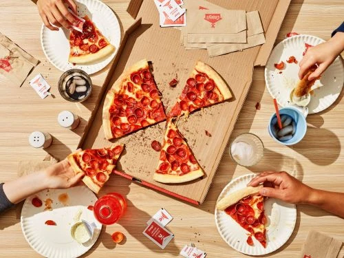 Overhead shot of group of people enjoying pepperoni pizza on paper plates with red beverages and condiments spread across a wooden table.
