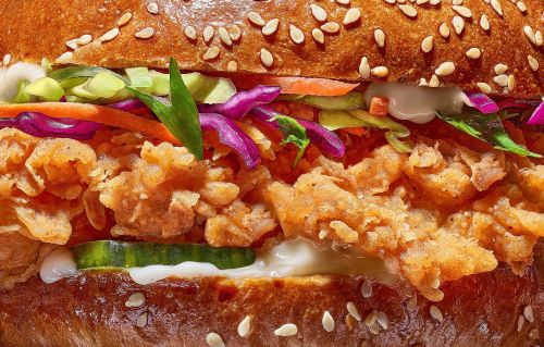 Close-up of a chicken sandwich featuring crispy fried chicken, shredded cabbage, sliced carrots, and sesame seed bun. Food photography.
