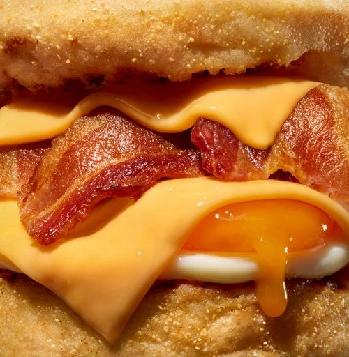 Close-up of a breakfast sandwich with melted cheese, bacon, and a fried egg with a dripping yolk between two halves of a bun.