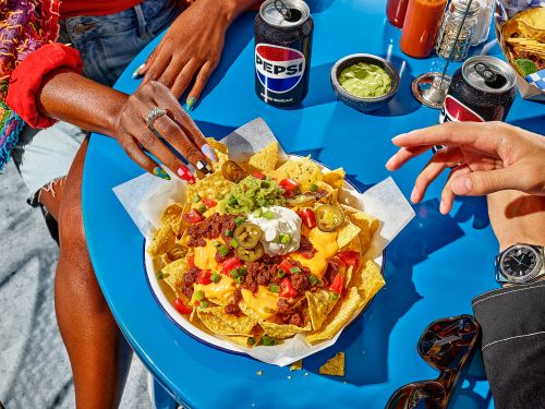 Overhead view of a vibrant blue table with a plate of loaded nachos, Pepsi cans, and hands reaching for the food, creating a social, culinary scene.