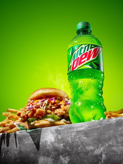 A refreshing food still life featuring a chicken sandwich and fries next to an ice-cold bottle of Mountain Dew soda on a gray surface.