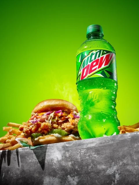 A refreshing food still life featuring a chicken sandwich and fries next to an ice-cold bottle of Mountain Dew soda on a gray surface.