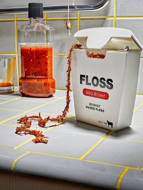 Novelty photo of a dental floss container labeled 'BBQ Brisket,' spilling strands of brisket-flavored floss onto a tiled countertop.
