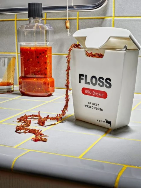 Novelty photo of a dental floss container labeled 'BBQ Brisket,' spilling strands of brisket-flavored floss onto a tiled countertop.