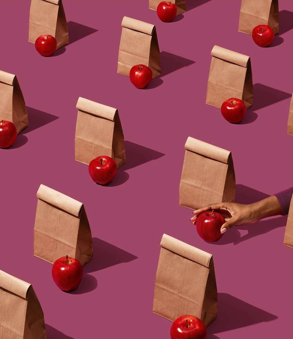 Still life pattern: red apples next to brown paper lunch bags on a purple background, one apple is being placed by a hand.