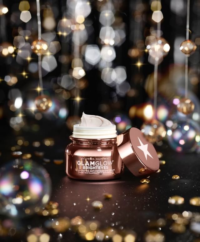 GlamGlow Brighteyes cream jar and lid sit on a black surface with bokeh lights for a beauty and skincare product shot.