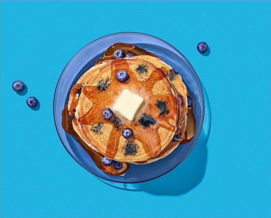 Overhead shot of pancakes with blueberries, butter, and syrup on a blue plate against a bright blue background, food photography.