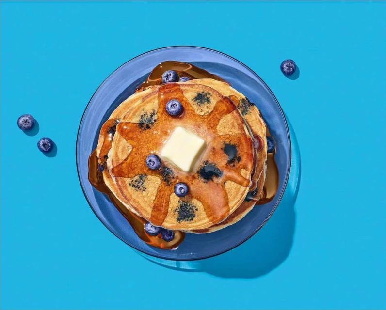 Overhead shot of pancakes with blueberries, butter, and syrup on a blue plate against a bright blue background, food photography.