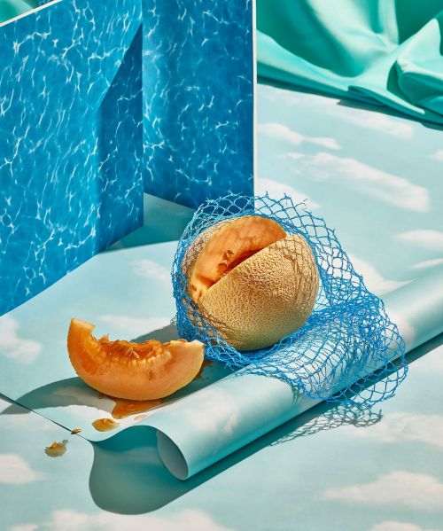 Studio shot of cantaloupe half in blue mesh, cantaloupe slice, and pool-water-patterned panels on a light blue surface, in a still life.