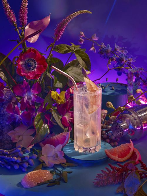 Cocktail drink on blue platform surrounded by vibrant flowers and greenery against a blue-purple background, cocktail shaker on the side.