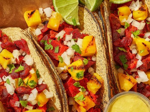 Three tacos al pastor with grilled pineapple, diced onion, cilantro, and lime wedges sit on a textured brown surface, close-up.