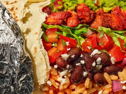 Close-up shot of a burrito open-faced with grilled meat, beans, rice, tomatoes, lettuce, cheese, and guacamole.