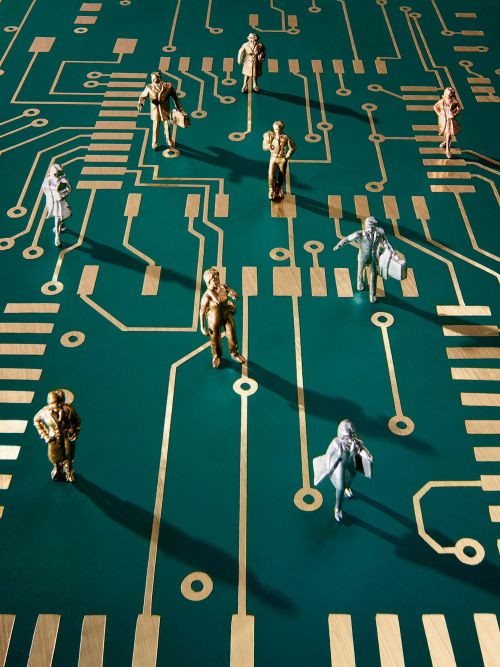 Figurines of people stand on a green circuit board with gold lines, resembling an electronic network or technology landscape.