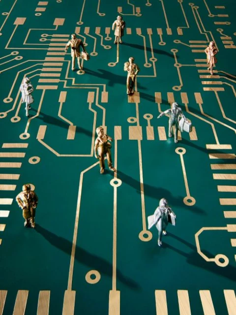 Figurines of people stand on a green circuit board with gold lines, resembling an electronic network or technology landscape.