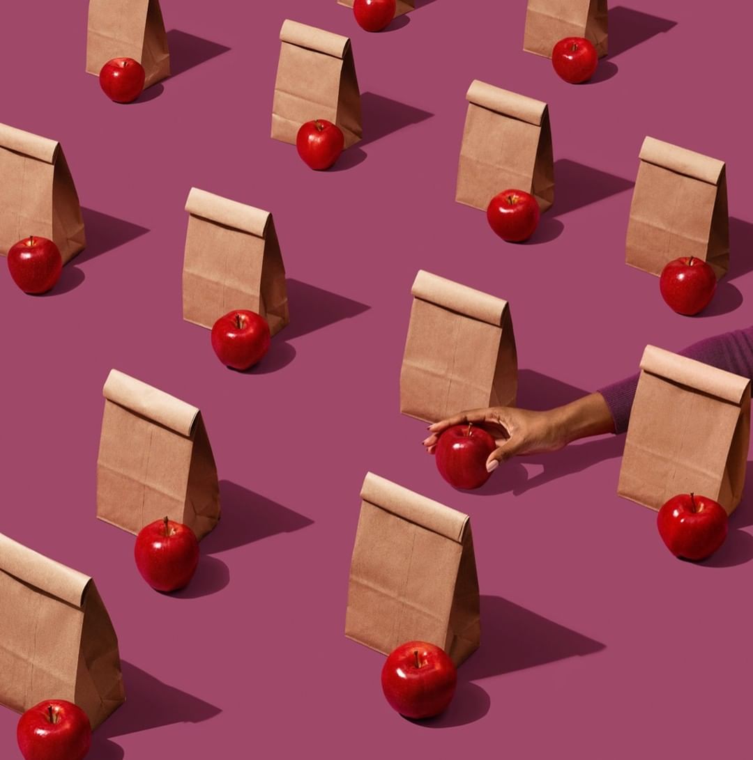 Overhead shot of multiple lunch bags and apples on a purple surface with one hand reaching for an apple, school lunch concept.