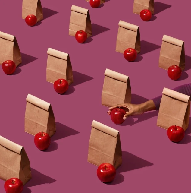 Overhead shot of multiple lunch bags and apples on a purple surface with one hand reaching for an apple, school lunch concept.