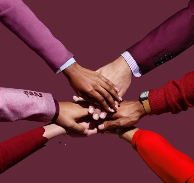 Diverse group of people stacks hands in a gesture of teamwork and unity, wearing suits and a watch against a solid maroon backdrop.