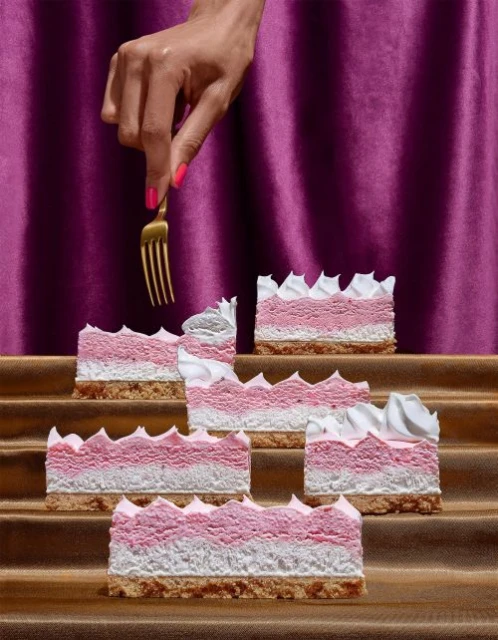 Stacked pink and white cakes are displayed with a golden fork held by a hand with painted nails against a purple curtain backdrop.
