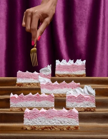 Stacked pink and white cakes are displayed with a golden fork held by a hand with painted nails against a purple curtain backdrop.