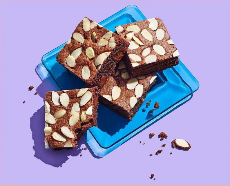Overhead shot of four almond brownies on a blue glass tray against a solid purple background, casting a soft shadow.